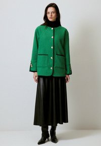 Jachetă din fleece texturat verde cu nasturi aurii și tiv negru, asortată cu o fustă din piele neagră și cizme negre până la genunchi.