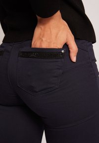 Jeans ajustados de color azul marino con una textura suave, que cuentan con un bolsillo trasero con borde de lentejuelas negras y un acento decorativo de rhinestones.