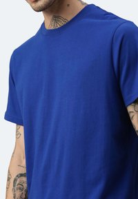 T-shirt blu in cotone con collo a giro, maniche corte e una texture liscia, dal fit relax, parzialmente coperto da un braccio tatuato.