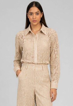 Femme portant une chemise en dentelle beige à manches longues et un pantalon taille haute en dentelle assorti, debout avec une main dans la poche, sur un fond uni.