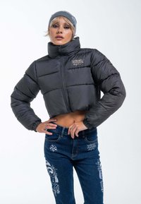 Gekürzte schwarze Daunenjacke mit hohem Kragen, Reißverschluss und elastischen Bündchen, kombiniert mit blauen Jeans mit weißen grafischen Designs.