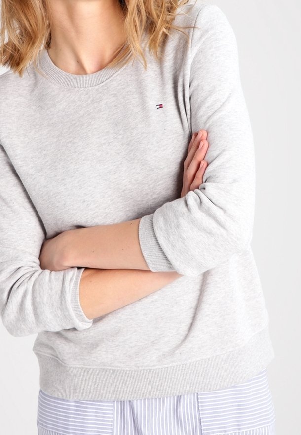 Femme aux cheveux blonds ondulés portant un sweat-shirt gris clair et un pantalon rayé, les bras croisés sur la poitrine sur un fond uni.