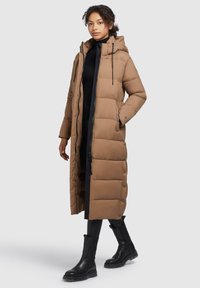 Manteau en duvet marron avec une capuche, design matelassé, fermeture éclair à l'avant, poches latérales et poignets élastiques, associé à des bottes noires et une robe.