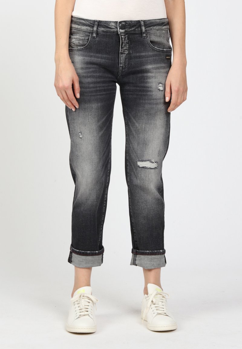 Nainen, jolla on ylösrullatut kuluneet mustat revityt farkut ja valkoiset lenkkarit, seisoo yksinkertaista taustaa vasten.