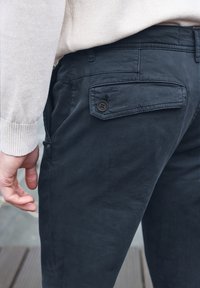 Námořnické modré kalhoty vyrobené z měkké tkaniny, s zadní kapsou a detaily knoflíku. Ruka je položená na bočním švu.