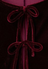 Chiusura di un indumento in velluto bordeaux con due fiocchi annodati, che presenta una texture liscia e una fodera interna coordinata di tonalità simile.