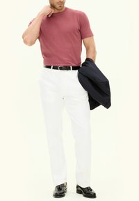 Maglietta girocollo bordeaux, pantaloni bianchi, scarpe in pelle nere e un blazer blu scuro appoggiato su un braccio. Design e vestibilità minimalisti.