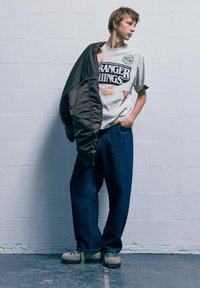 Młoda osoba w luźnych dżinsach i koszulce z motywem Stranger Things trzyma kurtkę na ramieniu, stojąc przy jasnoszarej ceglanej ścianie.