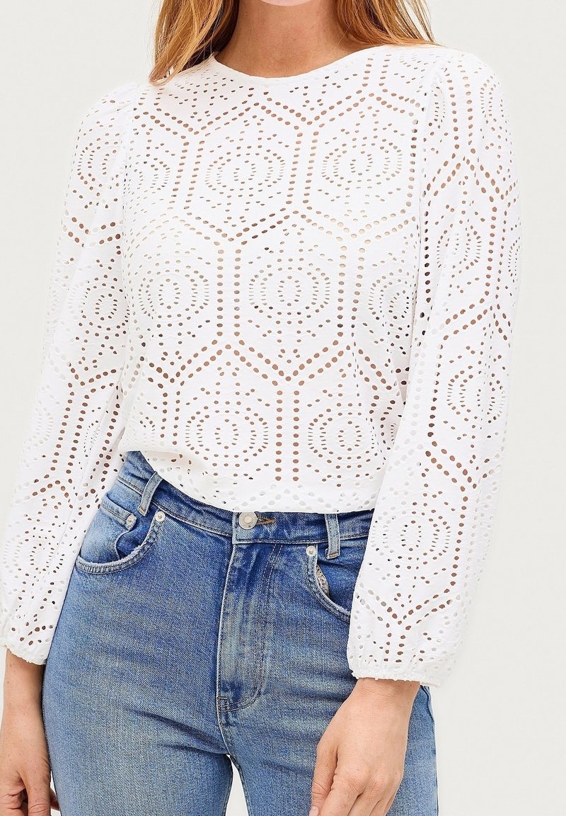 Weiße Bluse mit langen Ärmeln und geometrischem Lochmuster, kombiniert mit hochtaillierten blauen Jeans, von Hals bis Hüfte gezeigt.