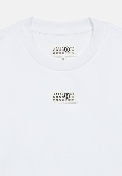 Bela bombažna majica z okrolim izrezom in majhno etiketo s številkami, pritrjeno na sredino izreza.
