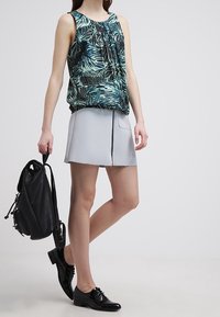 Top senza maniche con motivo floreale in blu e verde, abbinato a una gonna grigia chiara al ginocchio con zip frontale. Scarpe in pelle nere e zaino.