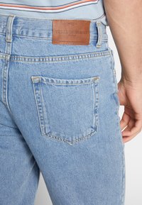 Jasnoniebieskie jeansy z denimu z skórzaną naszywką z tyłu, posiadające pięć kieszeni i widoczne detale przeszyć. Klasyczny krój.