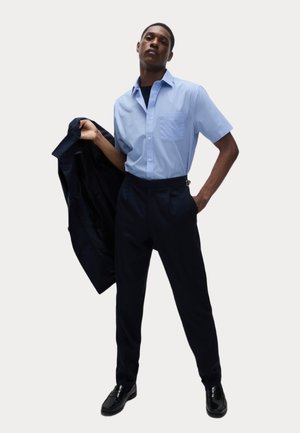 Jasnoniebieska koszula z krótkim rękawem z kieszonką na piersi, zestawiona z ciemnymi spodniami i czarnymi butami. Ciemna kurtka trzymana w prawej ręce.