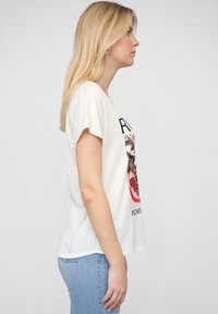 Valkoinen puuvillainen t-paita lyhyillä hihoilla, jossa on granaattiomenakuvio ja teksti. Yhdistetty vaaleansinisiin skinny-farkkuihin, sivuprofiilista.