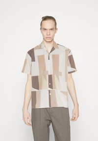 Camisa de manga corta, abotonada en beige claro, con patrones geométricos irregulares en marrón y crema. Tejido suave, corte relajado.