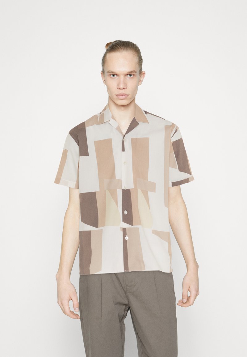 Camisa de manga corta, abotonada en beige claro, con patrones geométricos irregulares en marrón y crema. Tejido suave, corte relajado.