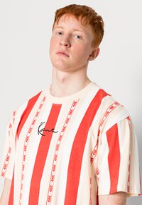 Rød og creme-stribet t-shirt med en afslappet pasform, der har et sort broderet logo og subtile pilmønstre langs striberne.