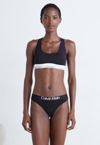Svart sport-bh i bomull med hvit Calvin Klein-logo på båndet, kombinert med svart bomulls-thong med logo foran. Enkel, tettsittende design.