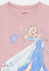 Roze t-shirt met een afbeelding van een personage met blond haar, gekleed in een blauwe jurk met een witte cape en sprankels, gelabeld "ELSA."