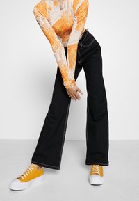 Mustat leveät farkut valkoisilla saumoilla yhdistettynä istuvaan pitkähiharaiseen paitaan, jossa on oranssi marmori-kuvio ja keltaiset lenkkarit.