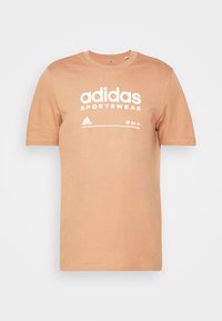 Světle hnědé bavlněné tričko s kulatým výstřihem, s bílým textem "adidas SPORTSWEAR" a logem. Hladká textura, krátké rukávy, uvolněný střih.