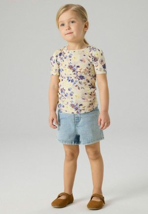 Ungt barn som står, iført en kortermet blomstret skjorte, lyseblå denimshorts og brune Mary Jane-sko mot en ensfarget bakgrunn.