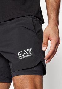 Juodi sportiniai šortai su lygiu paviršiumi, ant apatinės dalies turintys baltą EA7 Emporio Armani logotipą ir išskirtinės siūlės detales.