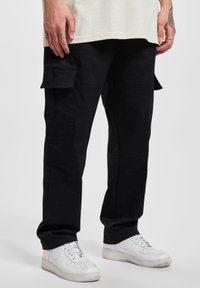 Pantalons cargo noirs avec poches latérales, fabriqués en tissu résistant. Présentent une coupe décontractée et des jambes droites, associés à des baskets blanches.