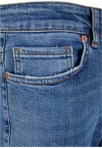 Jean en denim bleu clair avec une poche arrière. Le tissu présente un tissage classique et une texture subtile, avec des coutures orange contrastantes et un rivet en métal.