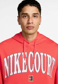 Felpa rossa con cappuccio e un grande grafica bianca "NIKECOURT", dotata di cappuccio con lacci e un piccolo patch multicolore del logo Nike sull'orlo.
