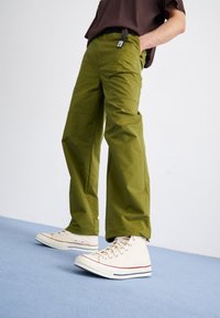 Olivgröna byxor med en avslappnad passform, fickor och en mjuk yta, tillsammans med beiga högsnörda sneakers med röda detaljer.