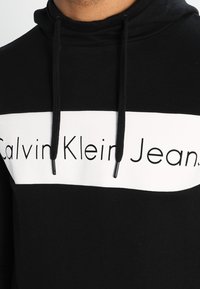 Felpa nera con cappuccio e una striscia orizzontale bianca che presenta il testo "Calvin Klein Jeans" in nero. Include lacci e un tessuto testurizzato.