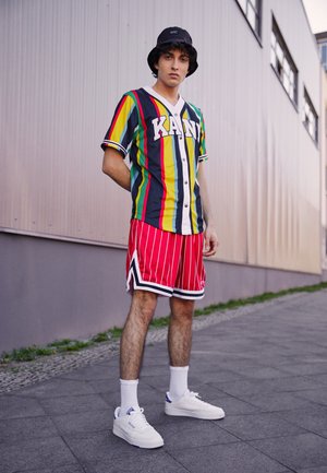 Jersey a rayas multicolor con el texto "KANE", pantalones cortos rojos a rayas, zapatillas blancas y sombrero de cubo negro; ambiente urbano al aire libre.