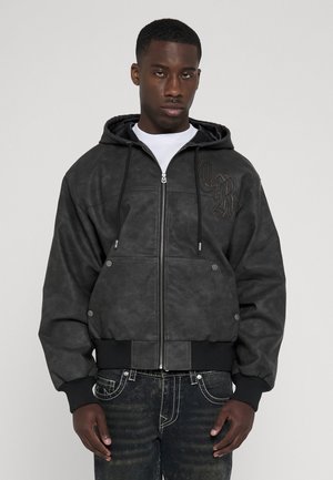 Giovane uomo che indossa una giacca nera con cappuccio e zip, con tasche frontali e jeans scuri con cuciture bianche, in piedi davanti a uno sfondo uniforme.