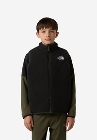 Fekete polár mellény magas gallérral és cipzáras lezárással, baloldali mellrészen logóval, sötétzöld hosszú ujjú ing fölött viselve.