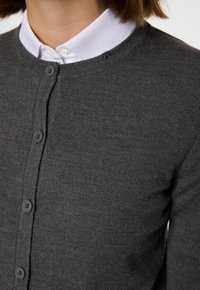 Grå stickad kofta med rund halsringning, knäppning fram och ribbade ärmar. Bärs över en vit skjorta med krage. Subtil textur med en mjuk finish.