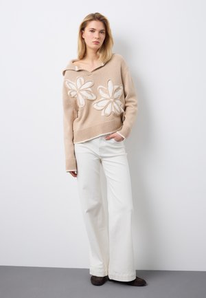 Giovane donna che indossa un maglione beige con grandi motivi floreali bianchi, pantaloni bianchi a gamba larga e scarpe scure, in piedi contro un muro bianco.