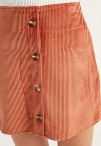 Jupe en velours côtelé rouille avec des cannelures verticales, taille haute et cinq boutons décoratifs marron sur le devant. Texture lisse, sans motifs.