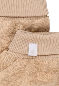Pull oversized beige en tissu texturé et duveteux, avec des poignets côtelés, et une petite étiquette blanche cousue sur le col.