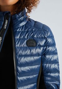 Personne portant une veste matelassée bleue brillante avec une fermeture éclair noire et un patch logo rond noir sur la poitrine.