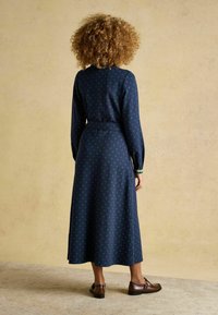 Garšs tumši zils kleita ar zaļiem punktiņiem, garām piedurknēm, jostu viduklī un izplūdušo apakšdaļu. Brūni apavi papildina izskatu.