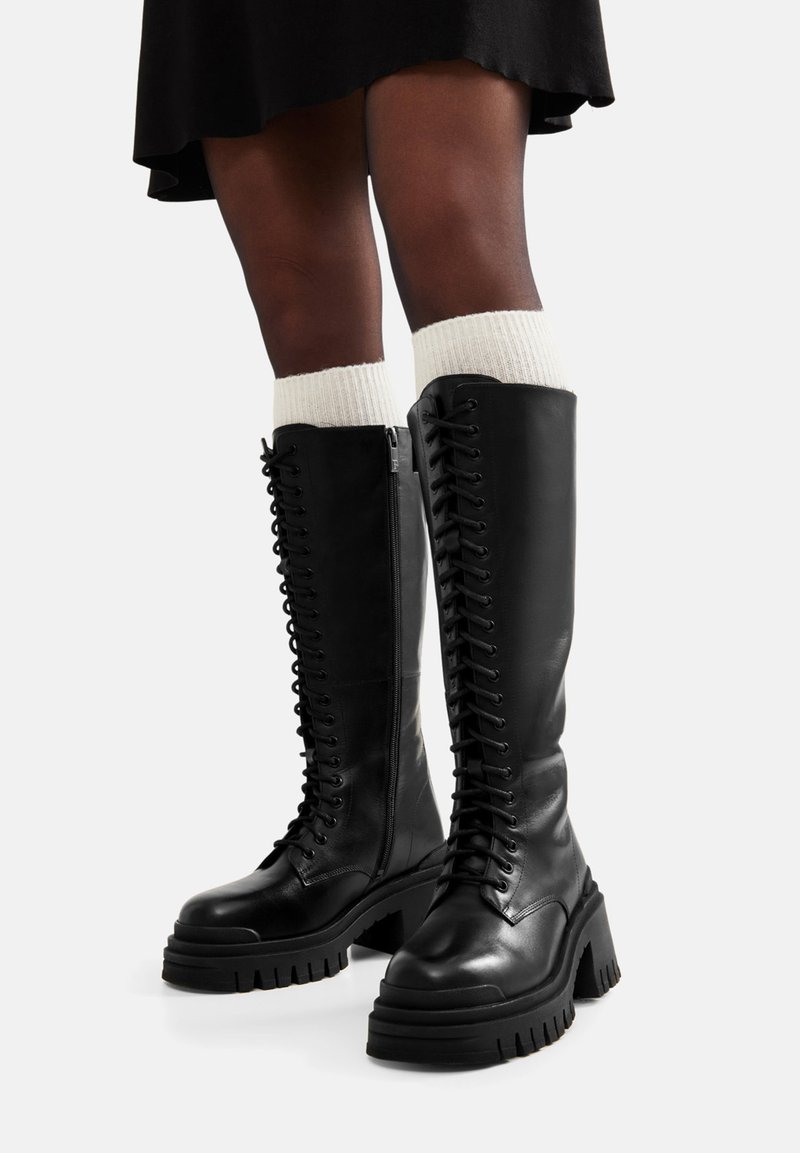 Czarne skórzane kozaki do kolan z grubą podeszwą na platformie, sznurowane z przodu i zapinane na boczny zamek, zestawione z kremowymi dzianinowymi skarpetami.