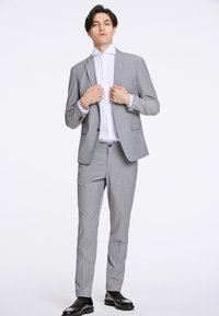 Grauer Nadelstreifenanzug mit strukturiertem Sakko und taillierten Hosen, kombiniert mit einem weißen Hemd mit Knöpfen. Glänzende schwarze Schuhe runden den Look ab.