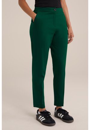 Frau trägt dunkelgrüne, maßgeschneiderte Hose und schwarze Adidas-Sneakers mit weißen Streifen, steht mit einer Hand in der Tasche.