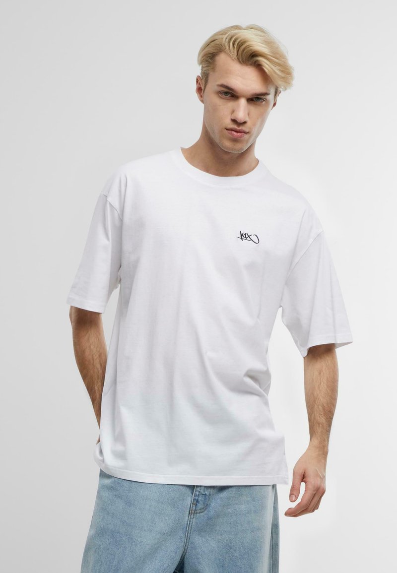 Hvid T-shirt med korte ærmer lavet af bomuld, med et lille sort logo på venstre bryst. Løs pasform, rund halsudskæring, glat tekstur.