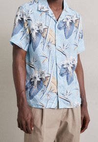 Camicetta a maniche corte realizzata in un tessuto leggero, con un motivo floreale blu e foglie. Ha una vestibilità rilassata e colletto con bottoni.