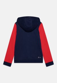 Sudadera azul marino con mangas rojas, hecha de algodón, que cuenta con una capucha ajustable, puños acanalados y un parche de logo cerca del dobladillo.