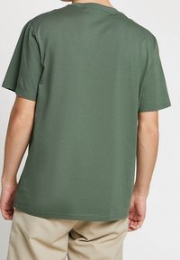 Person wearing plain green short-sleeve T-shirt and beige pants, shown from the back against a neutral background.