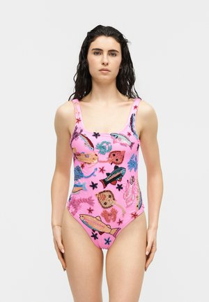 Γυναίκα με σκούρα σγουρά μαλλιά, φορώντας ένα ροζ μαγιό διακοσμημένο με πολύχρωμα ψάρια και στολίδια θαλάσσιων πλασμάτων, που στέκεται μπροστά σε ένα απλό φόντο.