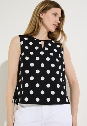 Frau trägt ärmelloses schwarzes Oberteil mit weißen Punkten und weiße Hose, schaut seitlich vor einfarbig hellem Hintergrund.